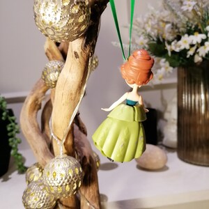 May include: A green and gold Christmas ornament featuring a cartoon princess with red hair. The ornament is hanging from a green ribbon on a wooden branch.