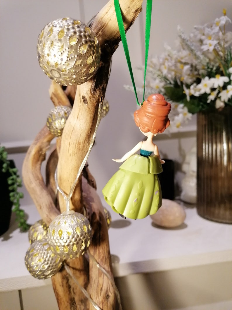 May include: A green and gold Christmas ornament featuring a cartoon princess with red hair. The ornament is hanging from a green ribbon on a wooden branch.