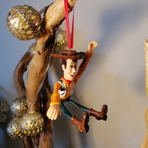 May include: A plastic toy figurine of Woody from the Toy Story franchise, wearing his signature cowboy hat and vest. He is hanging from a branch with a red ribbon.