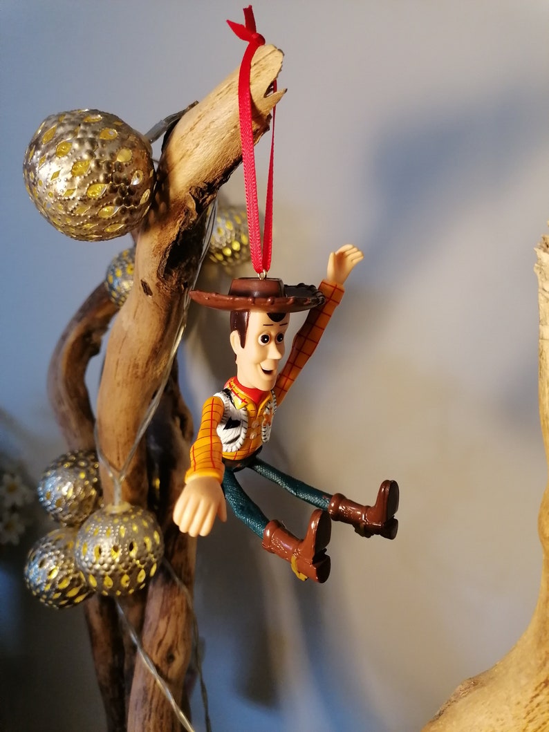 May include: A plastic toy figurine of Woody from the Toy Story franchise, wearing his signature cowboy hat and vest. He is hanging from a branch with a red ribbon.