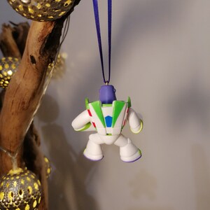 May include: A white, green and purple Buzz Lightyear Christmas ornament hanging from a purple ribbon. The ornament is a small plastic figure of Buzz Lightyear from the Toy Story films.