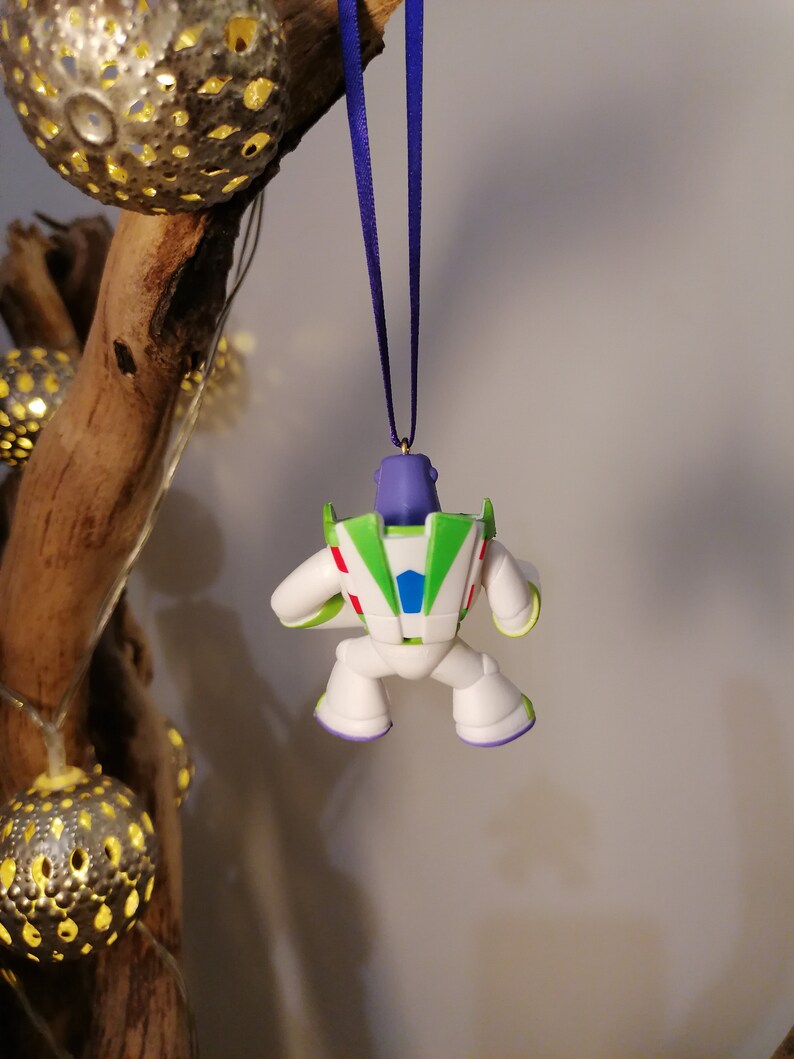 May include: A white, green and purple Buzz Lightyear Christmas ornament hanging from a purple ribbon. The ornament is a small plastic figure of Buzz Lightyear from the Toy Story films.