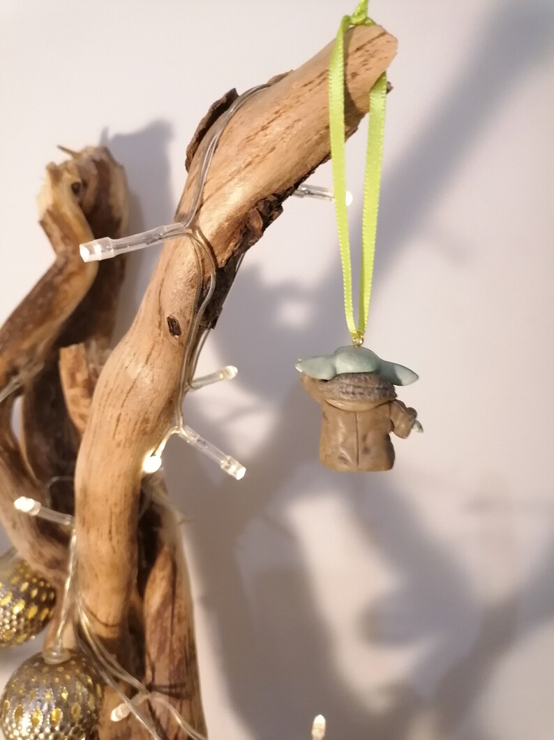 Pu&ograve; includere: Una statuetta verde e marrone di un personaggio del franchise di Star Wars, appesa a un nastro verde su un ramo di legno galleggiante. La statuetta indossa una tunica marrone e ha una testa verde.
