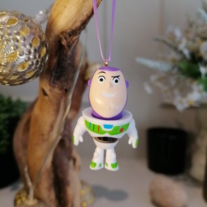 Puede incluir: Un adorno de pl&aacute;stico morado y blanco de Buzz Lightyear de la franquicia Toy Story. El adorno est&aacute; colgado de una cinta morada.
