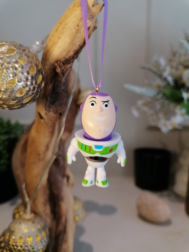 Puede incluir: Un adorno de pl&aacute;stico morado y blanco de Buzz Lightyear de la franquicia Toy Story. El adorno est&aacute; colgado de una cinta morada.