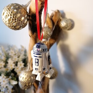 K&ouml;nnte beinhalten: Ein wei&szlig;er und blauer R2-D2 Star Wars Charakter Anh&auml;nger, der an einem roten Band an einem Holzast h&auml;ngt. Der Anh&auml;nger ist eine Weihnachtsbaumdekoration.