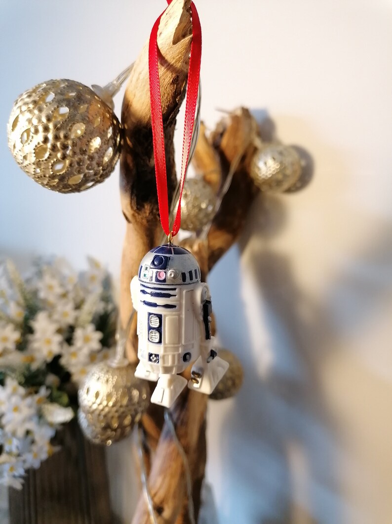 K&ouml;nnte beinhalten: Ein wei&szlig;er und blauer R2-D2 Star Wars Charakter Anh&auml;nger, der an einem roten Band an einem Holzast h&auml;ngt. Der Anh&auml;nger ist eine Weihnachtsbaumdekoration.
