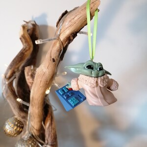 May include: A green and brown Christmas ornament featuring a character from the Star Wars franchise, The Mandalorian. The ornament is wearing a white coat and holding a blue device. The ornament is hanging from a green ribbon on a wooden branch.