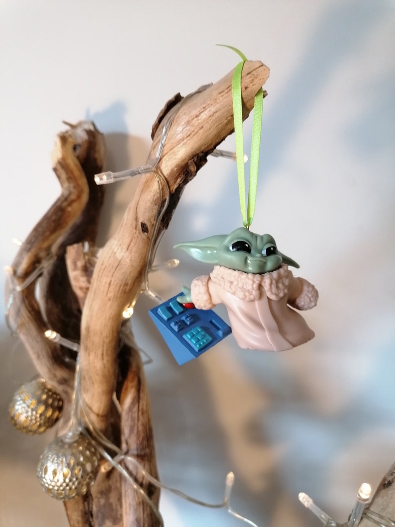 May include: A green and brown Christmas ornament featuring a character from the Star Wars franchise, The Mandalorian. The ornament is wearing a white coat and holding a blue device. The ornament is hanging from a green ribbon on a wooden branch.