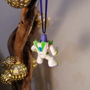 May include: A white, green and purple plastic ornament of Buzz Lightyear from the Toy Story franchise. The ornament is hanging from a blue ribbon.