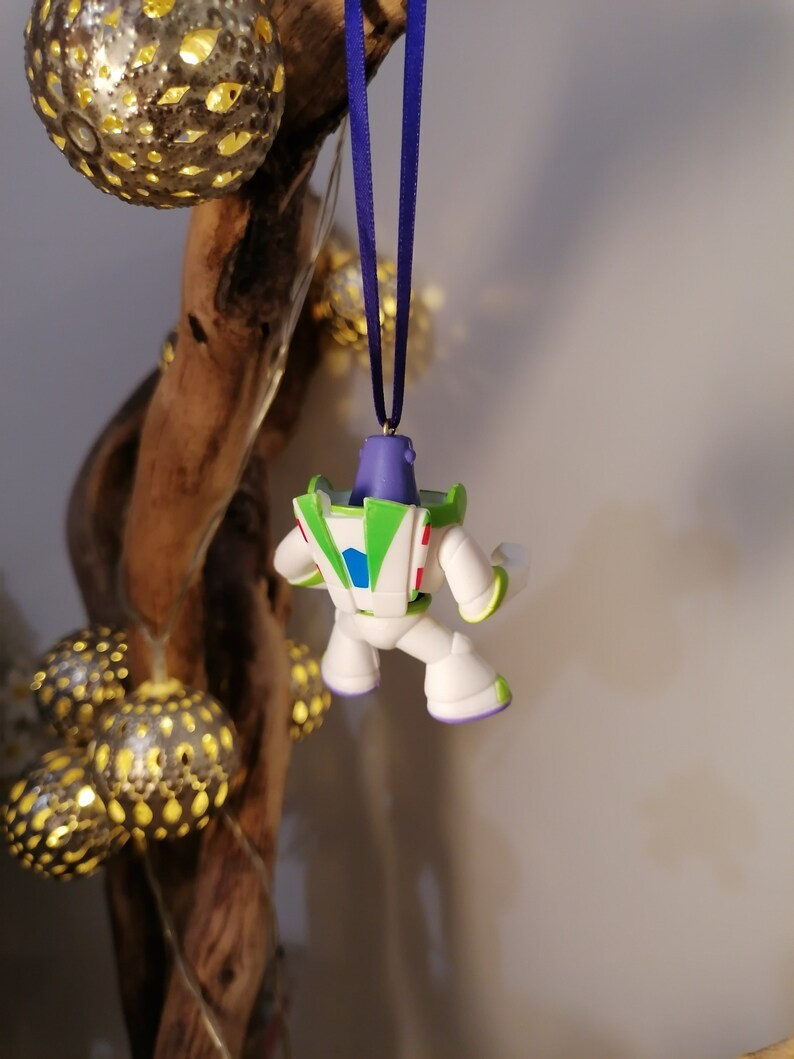 May include: A white, green and purple plastic ornament of Buzz Lightyear from the Toy Story franchise. The ornament is hanging from a blue ribbon.