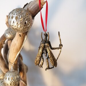 May include: A gold-coloured insect ornament with wings and a bow and arrow. The ornament is hanging from a red ribbon.
