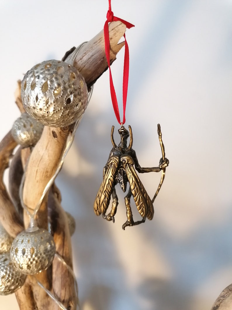 May include: A gold-coloured insect ornament with wings and a bow and arrow. The ornament is hanging from a red ribbon.