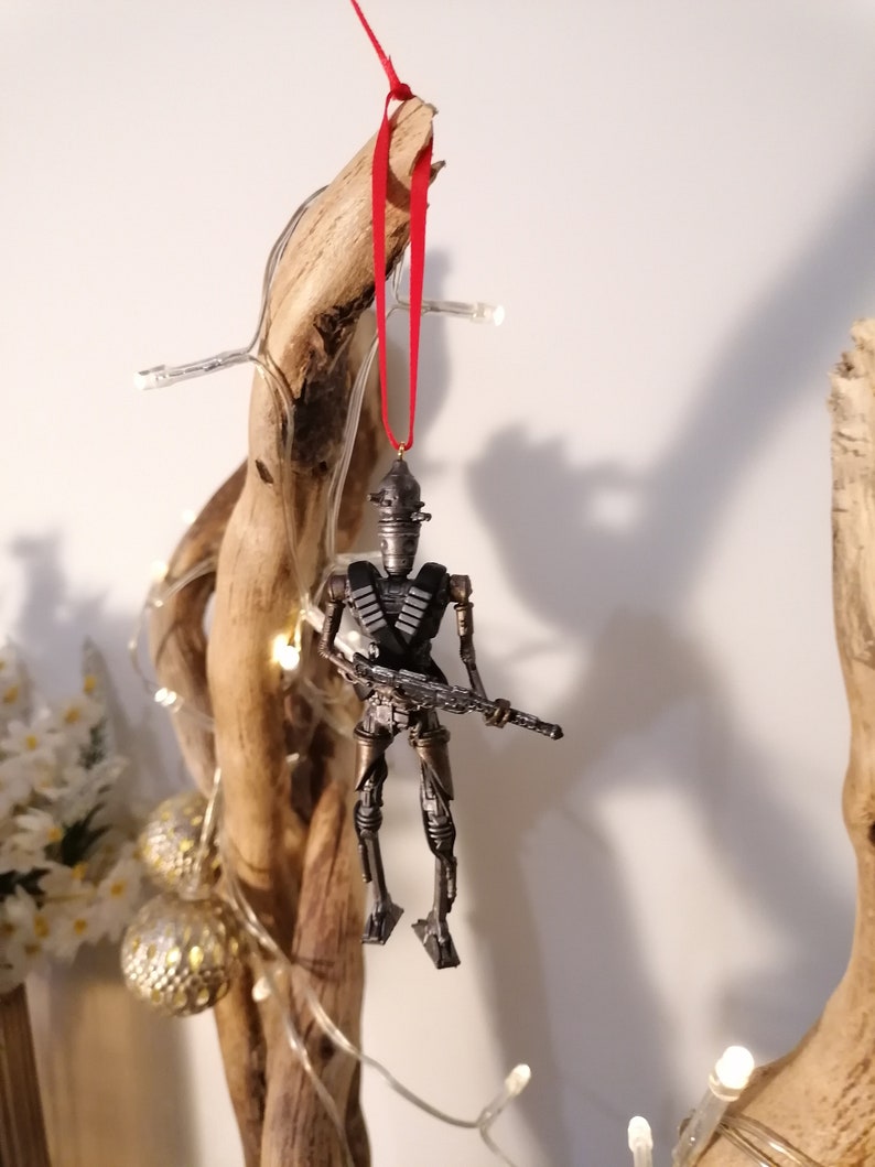 Puede incluir: Un adorno de robot de color plateado con una pistola colgando de una cinta roja en una rama de madera. El robot lleva un bandolera y tiene un dise&ntilde;o detallado.