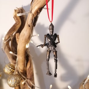 Puede incluir: Un adorno de robot plateado y negro colgado de una rama con una cinta roja. El robot sostiene una pistola y tiene una cruz atada en el pecho.