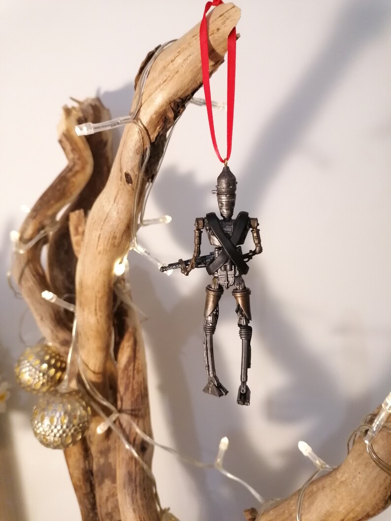 Puede incluir: Un adorno de robot plateado y negro colgado de una rama con una cinta roja. El robot sostiene una pistola y tiene una cruz atada en el pecho.