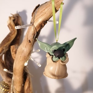 May include: Green and brown Christmas ornament shaped like a character from the Star Wars franchise, hanging from a green ribbon on a branch.