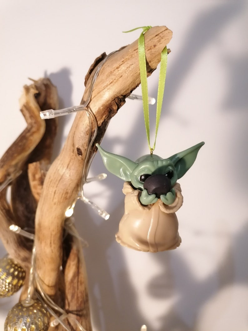 May include: Green and brown Christmas ornament shaped like a character from the Star Wars franchise, hanging from a green ribbon on a branch.