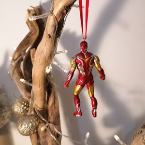 May include: Red and gold Iron Man Christmas ornament hanging from a branch with white lights. The ornament is a detailed figurine of Iron Man in a flying pose.