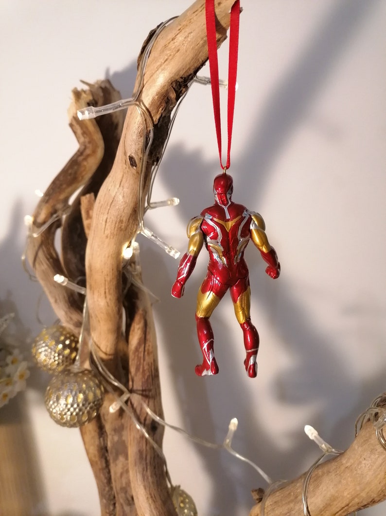 May include: Red and gold Iron Man Christmas ornament hanging from a branch with white lights. The ornament is a detailed figurine of Iron Man in a flying pose.