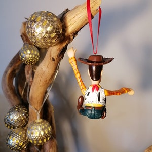 May include: A red string hangs from a branch with a plastic toy figurine of Woody from Toy Story. Woody is wearing his signature cowboy hat, vest, and jeans. He is holding onto the branch with one hand and his other arm is outstretched.