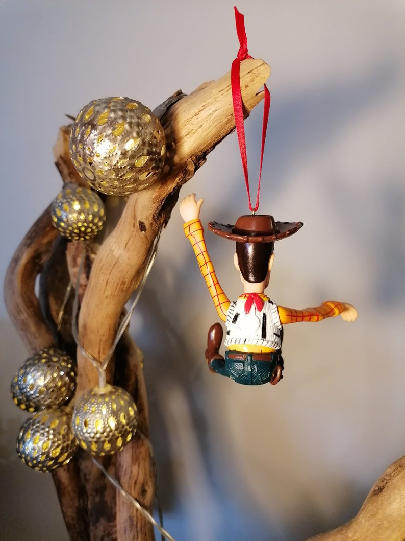 May include: A red string hangs from a branch with a plastic toy figurine of Woody from Toy Story. Woody is wearing his signature cowboy hat, vest, and jeans. He is holding onto the branch with one hand and his other arm is outstretched.