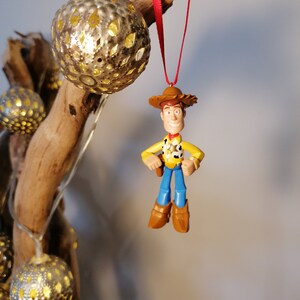 May include: A red string hangs from a wooden branch with a gold ornament and a plastic toy figurine of Woody from Toy Story. Woody is wearing his signature cowboy hat, vest, and jeans.