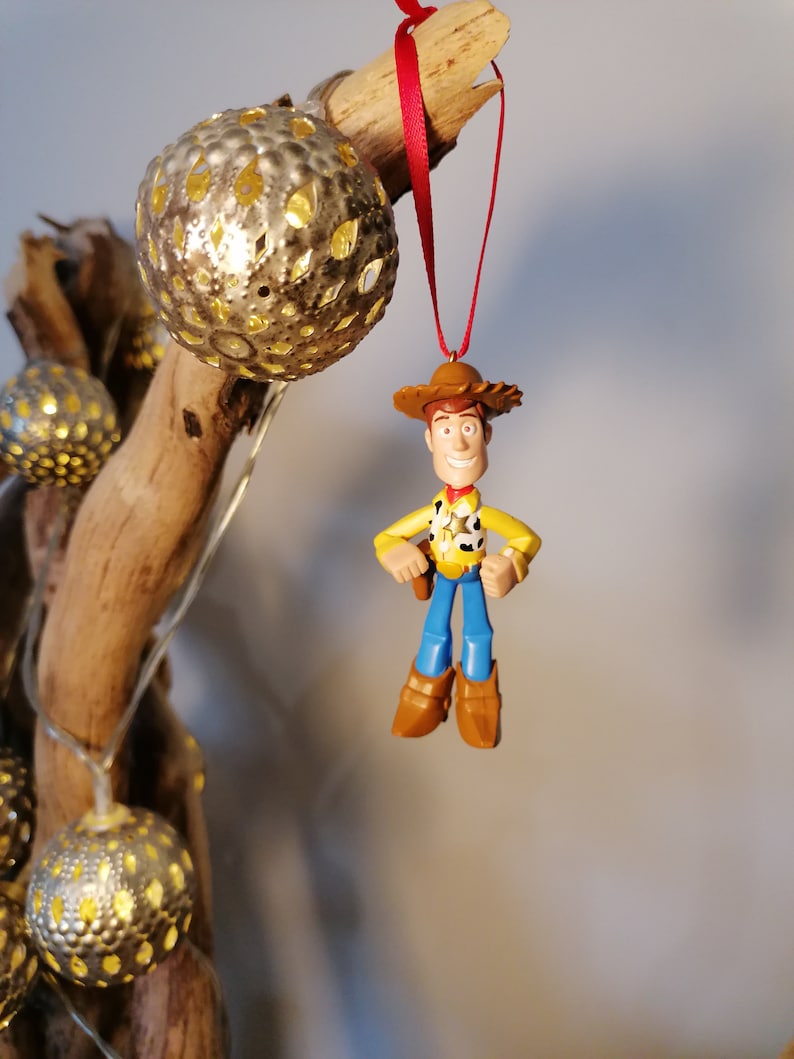 May include: A red string hangs from a wooden branch with a gold ornament and a plastic toy figurine of Woody from Toy Story. Woody is wearing his signature cowboy hat, vest, and jeans.