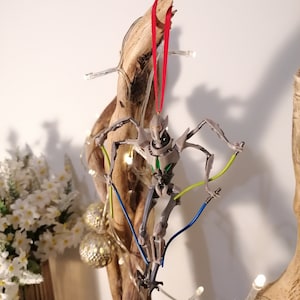May include: A gray and green Star Wars General Grievous character ornament hanging from a branch with a red ribbon. The ornament has a light blue and yellow wire attached to it.