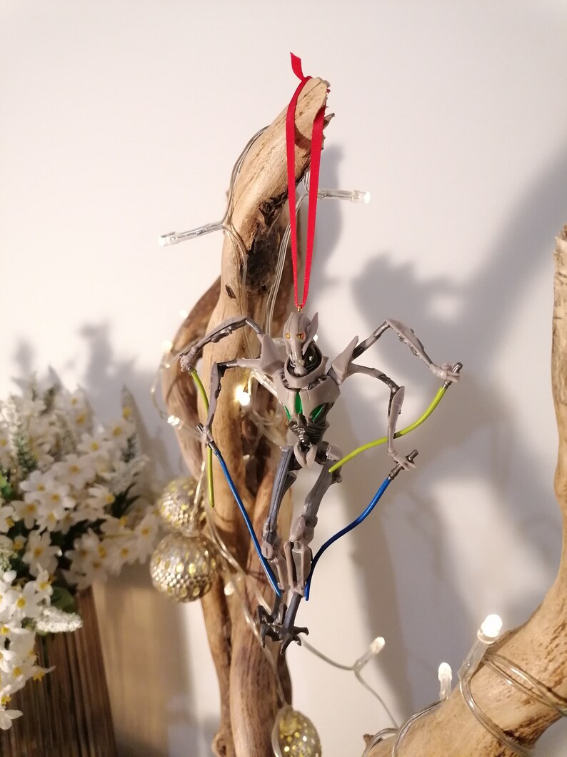 May include: A gray and green Star Wars General Grievous character ornament hanging from a branch with a red ribbon. The ornament has a light blue and yellow wire attached to it.