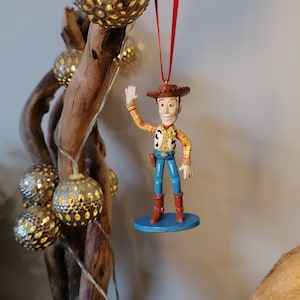 May include: A red and white cowboy figurine ornament of Woody from Toy Story, with a blue base, hanging from a red ribbon on a branch.