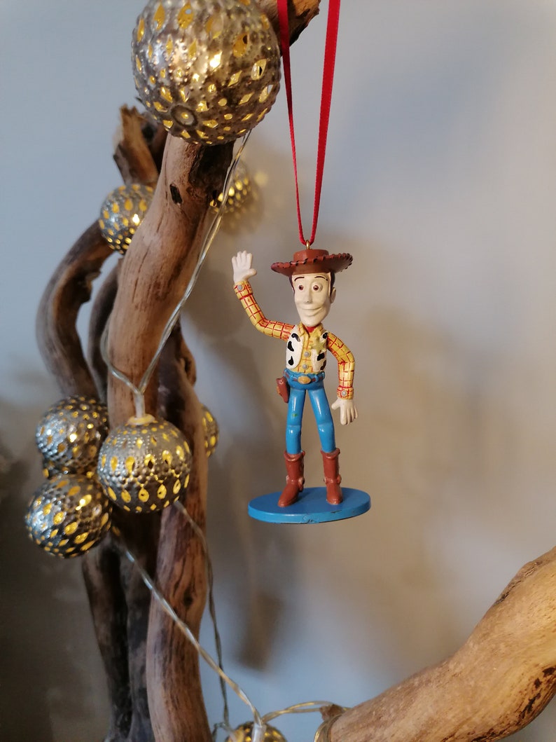 May include: A red and white cowboy figurine ornament of Woody from Toy Story, with a blue base, hanging from a red ribbon on a branch.
