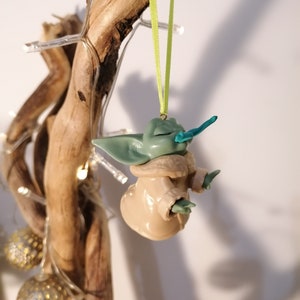 May include: A green and brown handmade ornament of a popular fictional character, hanging from a green ribbon on a branch.
