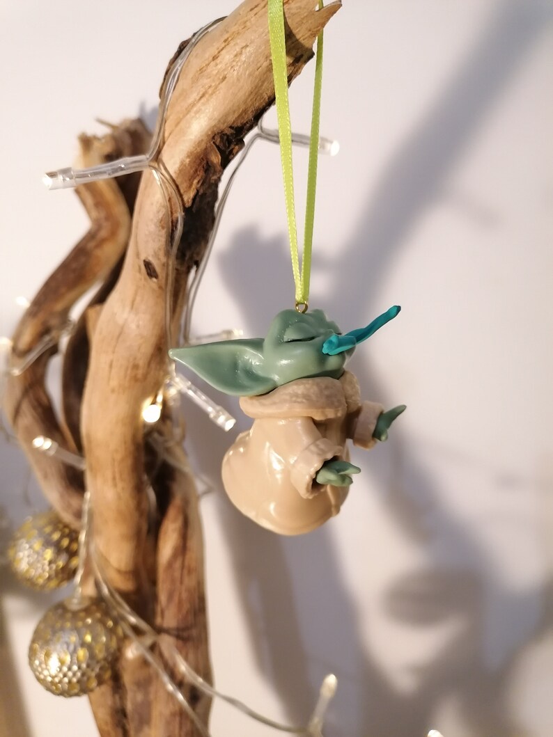 May include: A green and brown handmade ornament of a popular fictional character, hanging from a green ribbon on a branch.