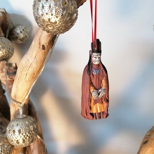 May include: A red string ornament with a detailed figurine of a character from a fantasy movie. The character is wearing a brown robe and a crown. The ornament is hanging from a branch with silver decorative lights.