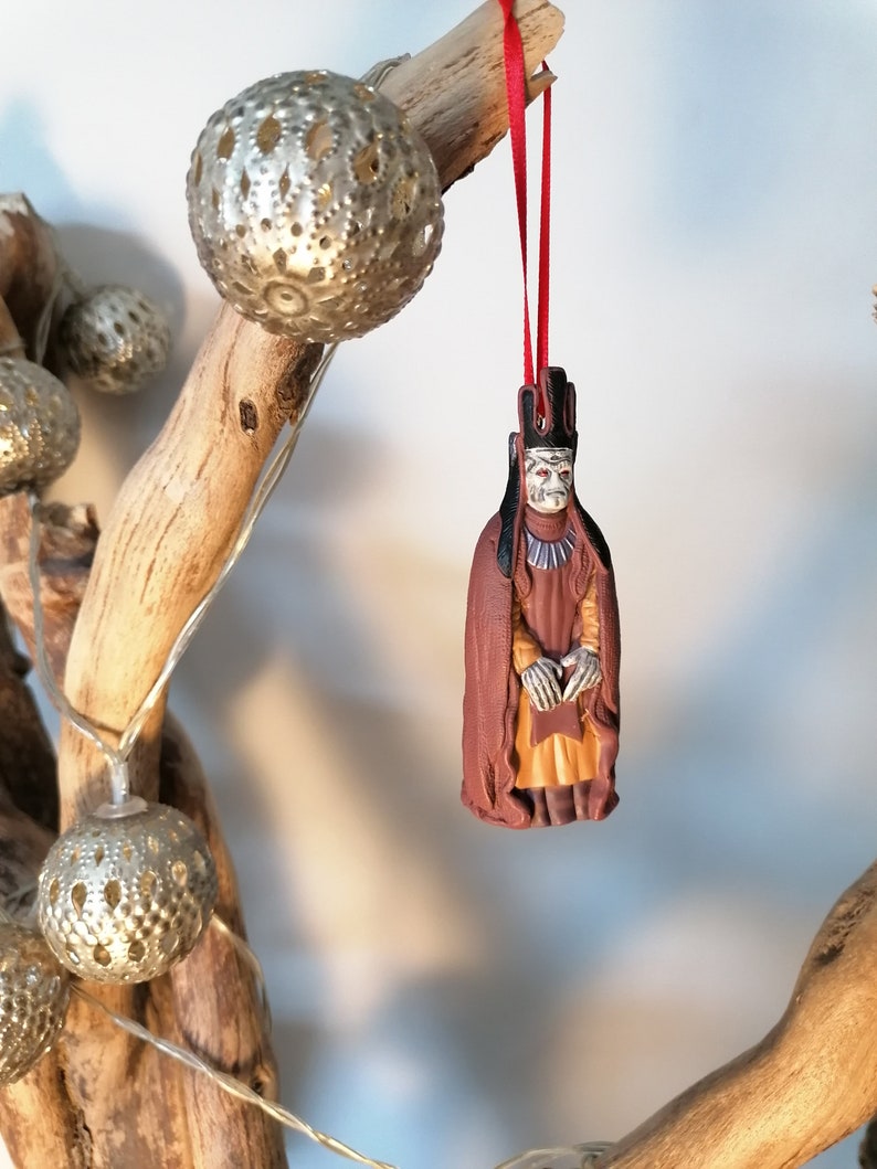 May include: A red string ornament with a detailed figurine of a character from a fantasy movie. The character is wearing a brown robe and a crown. The ornament is hanging from a branch with silver decorative lights.