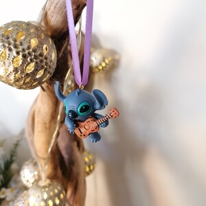 Op de afbeelding: Een blauwe Stitch-figuur ornament dat een bruine ukelele bespeelt, hangend aan een paars lint.