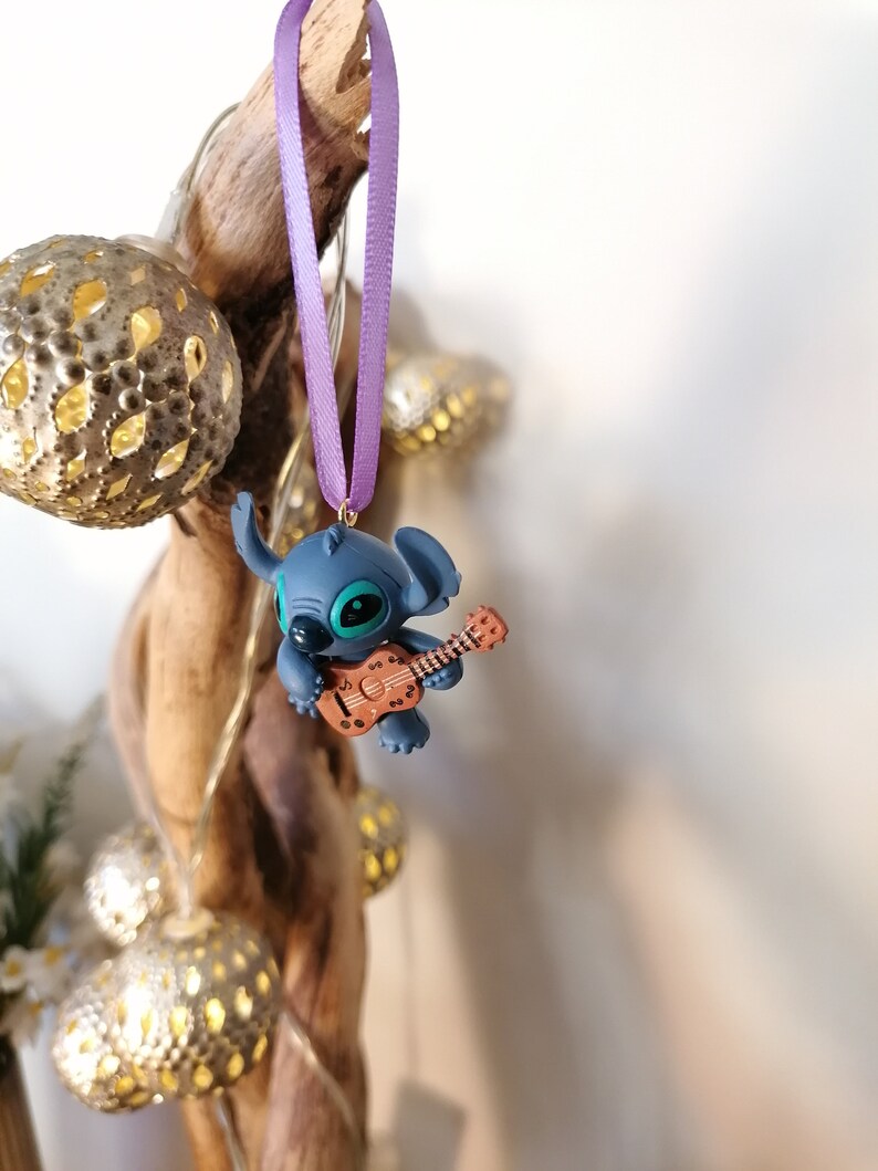 Op de afbeelding: Een blauwe Stitch-figuur ornament dat een bruine ukelele bespeelt, hangend aan een paars lint.