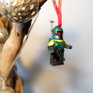 May include: A green and brown Lego Star Wars Boba Fett figure with a red ribbon hanging from a wooden branch. The figure is holding two blasters.