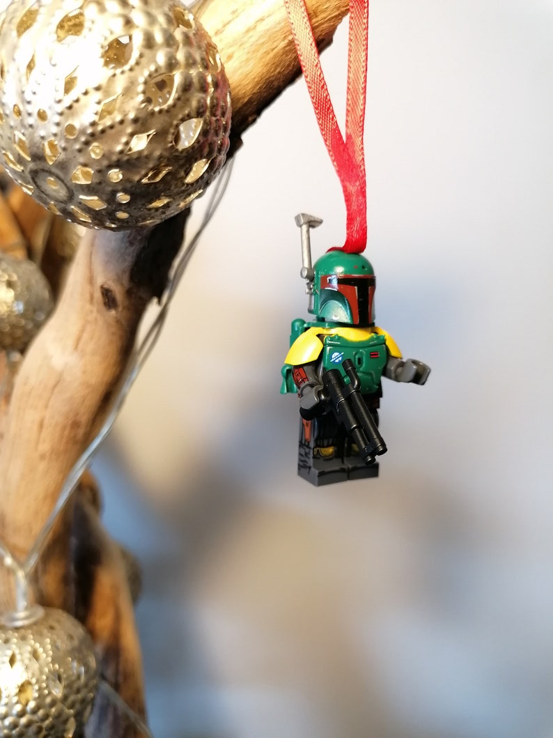May include: A green and brown Lego Star Wars Boba Fett figure with a red ribbon hanging from a wooden branch. The figure is holding two blasters.
