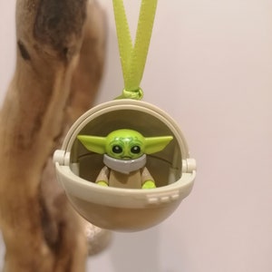 May include: A green and tan Baby Yoda figure inside a tan pod, hanging from a light green ribbon. The figure has large eyes and ears. The pod is round and has a hinged opening. The background is a light color.