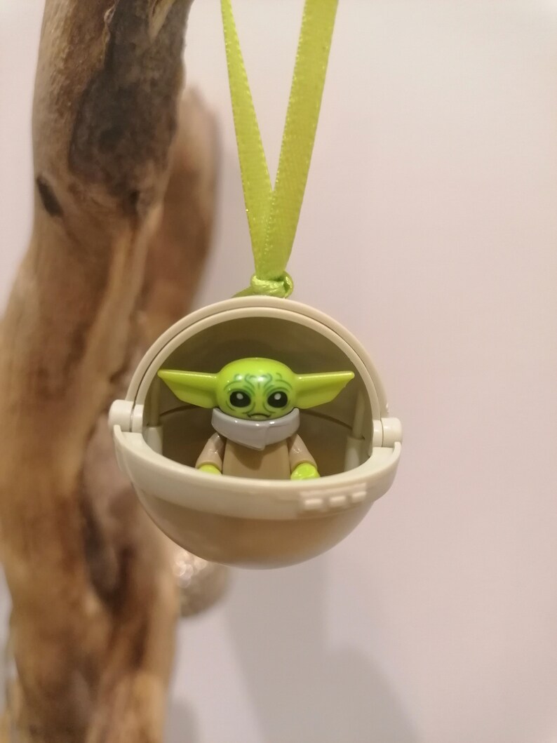 May include: A green and tan Baby Yoda figure inside a tan pod, hanging from a light green ribbon. The figure has large eyes and ears. The pod is round and has a hinged opening. The background is a light color.