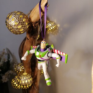 May include: A purple and white plastic toy figurine of Buzz Lightyear from the Toy Story movie franchise hanging from a purple ribbon on a wooden branch. The figurine is wearing a green and white spacesuit with a red and white stripe on the back. The figurine is holding a purple and white laser gun.