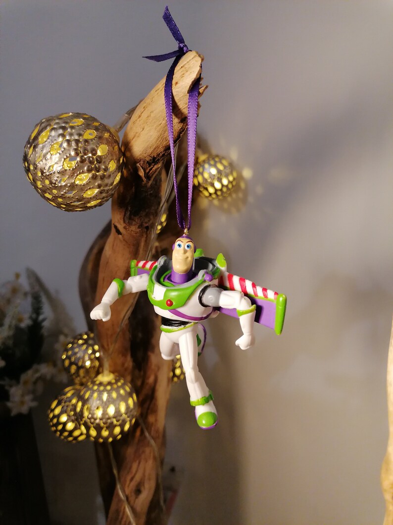 May include: A purple and white plastic toy figurine of Buzz Lightyear from the Toy Story movie franchise hanging from a purple ribbon on a wooden branch. The figurine is wearing a green and white spacesuit with a red and white stripe on the back. The figurine is holding a purple and white laser gun.