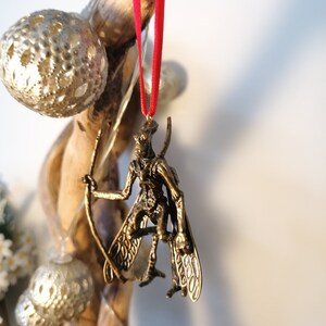 May include: A gold-coloured ornament of a winged creature with a long, thin tail. The ornament is hanging from a red ribbon.