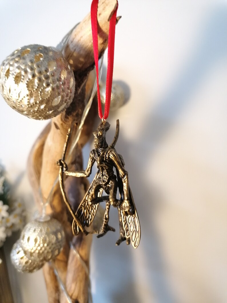 May include: A gold-coloured ornament of a winged creature with a long, thin tail. The ornament is hanging from a red ribbon.