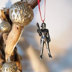 May include: A silver-coloured ornament of a female figure holding a weapon. The ornament is hanging from a red ribbon.