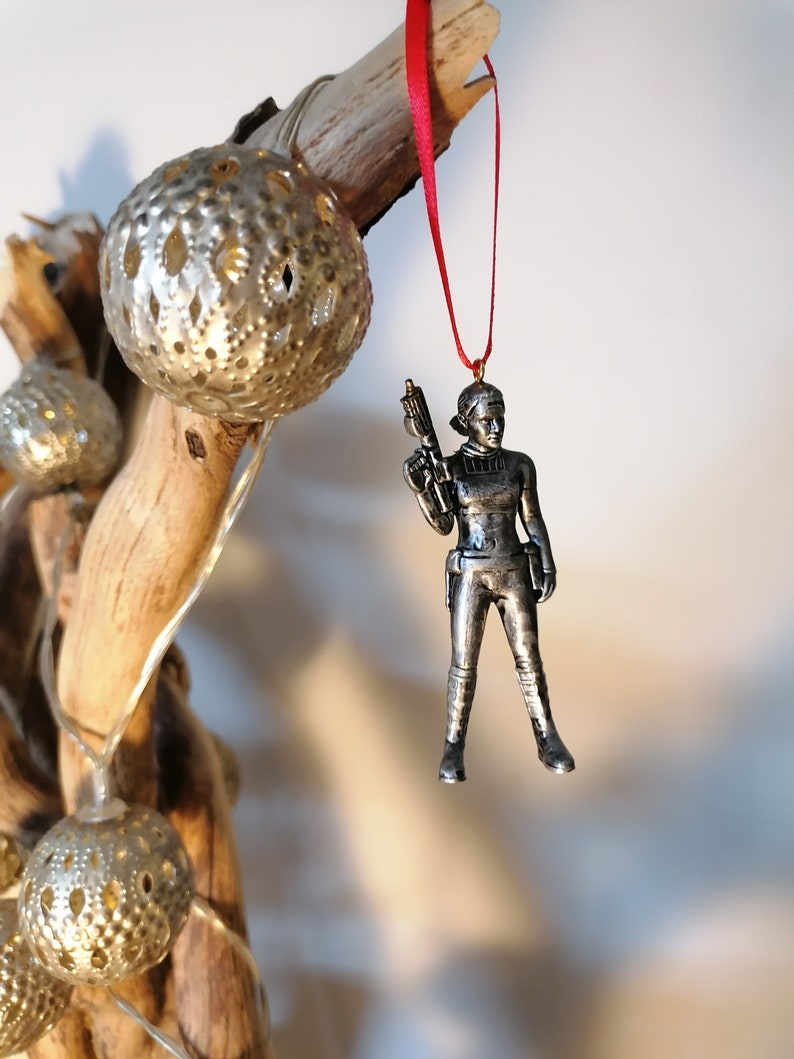 May include: A silver-coloured ornament of a female figure holding a weapon. The ornament is hanging from a red ribbon.