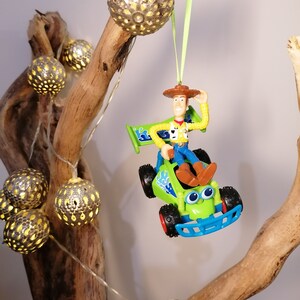May include: A green toy car with a blue base and a figure of Woody from Toy Story driving it. The car is hanging from a branch with a string of lights. The lights are silver and have a cut-out design.