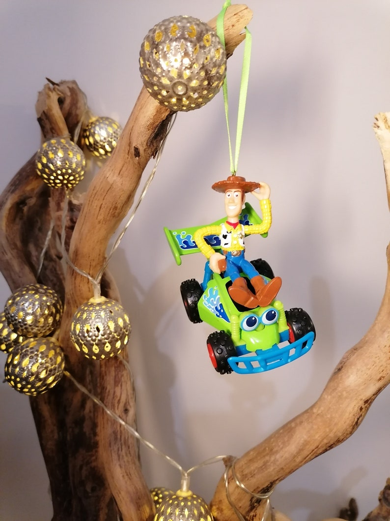 May include: A green toy car with a blue base and a figure of Woody from Toy Story driving it. The car is hanging from a branch with a string of lights. The lights are silver and have a cut-out design.